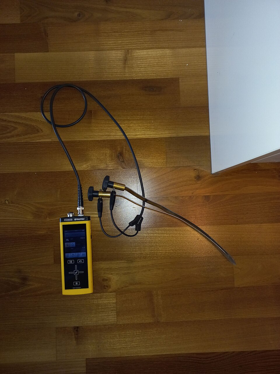 A pin-type moisture meter with two corded probe electrodes lying on a wood floor