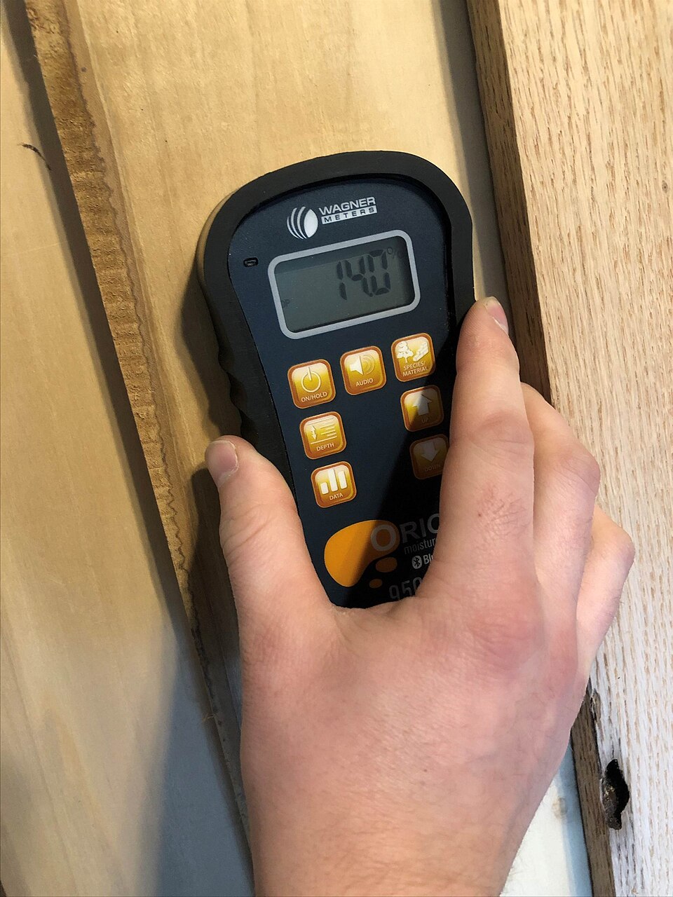 A pinless moisture meter held flat against a rough-sawn board, its display reading 14.0 percent