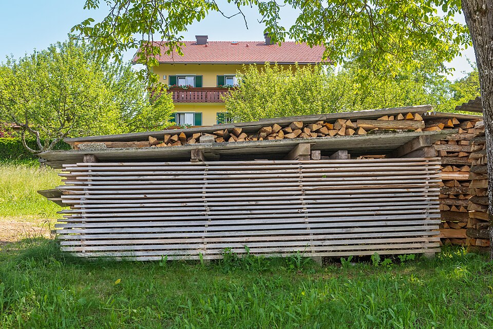 A tall stack of rough-sawn boards separated by thin sticker strips, air-drying under a simple plank roof